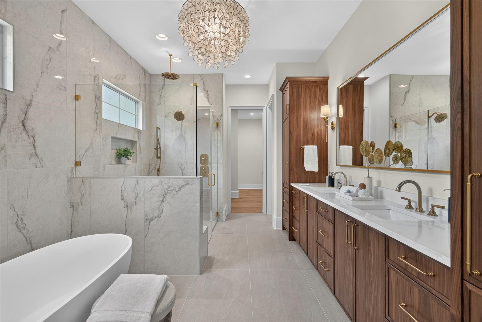 Primary bath at Heather wrapped in book-matched marble slab with freestanding tub, glass shower, and walnut double vanity beneath a capiz-shell chandelier — Heather by Stillman Properties Charlotte NC