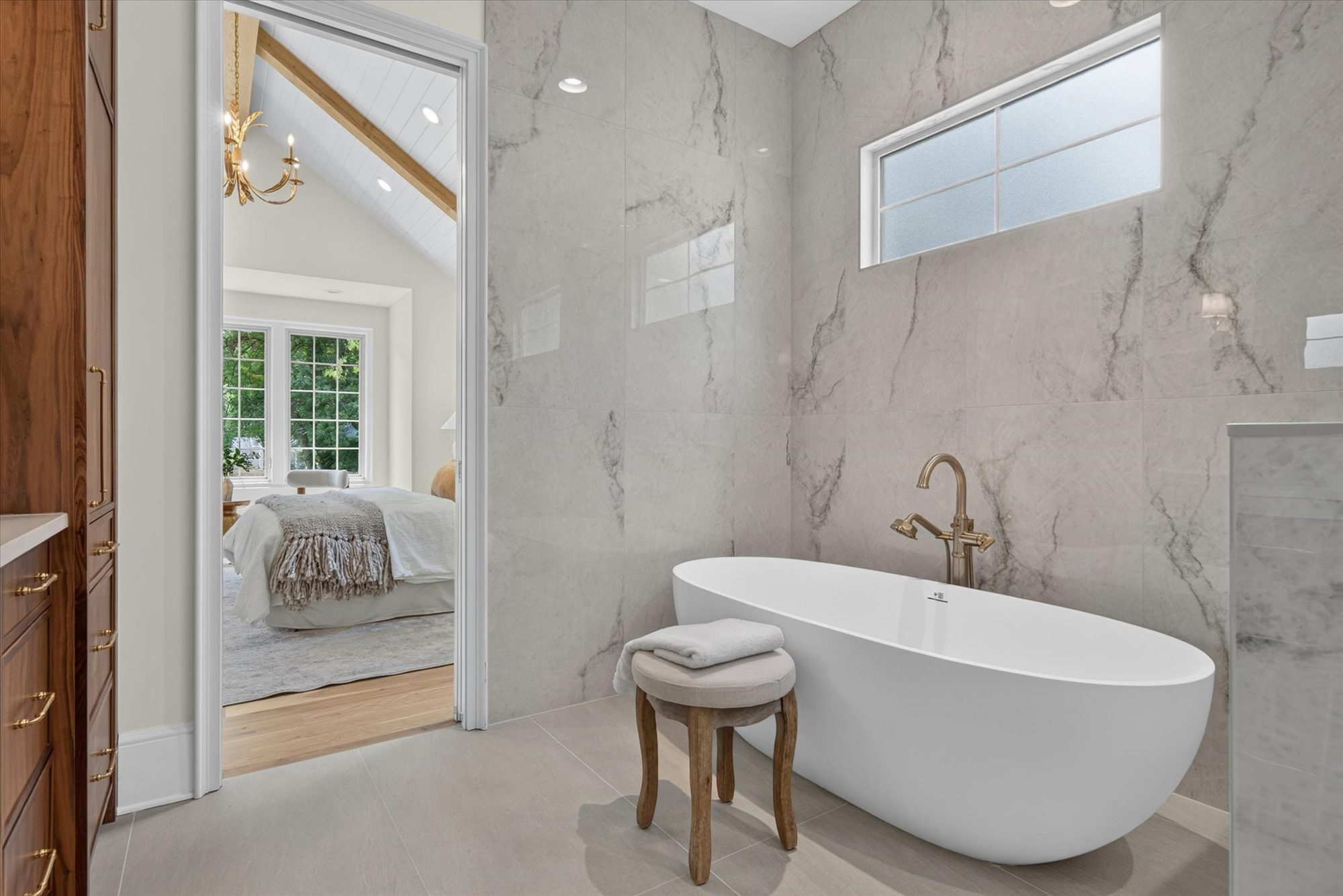 Freestanding soaking tub at Heather set against a full-height marble slab wall below a clerestory window, with the vaulted primary bedroom beyond — Heather by Stillman Properties Charlotte NC